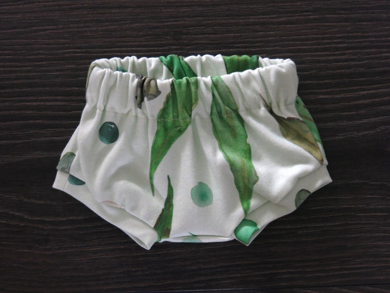May include: White baby bloomers with an elastic waistband, featuring a watercolor print of green leaves and dots. The fabric is soft and the design is gender-neutral.