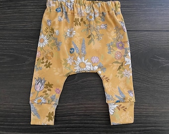 May include: Mustard yellow pants featuring an all-over floral print with white daisies, purple lavender, and various green foliage. The pants have an elasticized waistband and fitted cuffs at the ankles, providing a comfortable and secure fit.