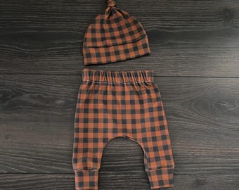 May include: A two-piece set consisting of a knotted beanie hat and matching pants, both featuring a dark brown and burnt orange buffalo plaid check pattern. The pants have an elasticized waistband and cuffed ankles for a secure fit.