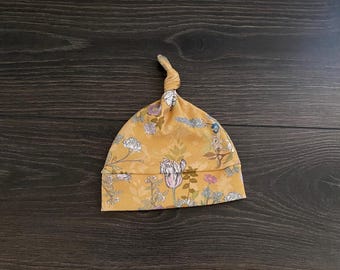 May include: A mustard yellow baby hat featuring a knotted top and a folded brim. The fabric is decorated with a botanical print consisting of various flowers, leaves, and stems in shades of white, pink, light blue, and muted green throughout.