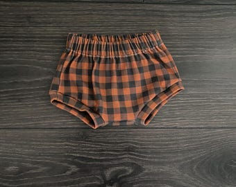 May include: A pair of baby bloomers featuring a dark brown and black buffalo plaid check pattern. The garment has an elasticized waistband and elasticized leg openings, providing a gathered, ruffled appearance around the waist and thighs.