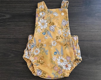 May include: A mustard yellow romper featuring a floral pattern with white daisies, blue wildflowers, and muted pink blossoms. The garment has two shoulder straps, a square neckline with two white buttons, and elasticized leg openings for a gathered fit.