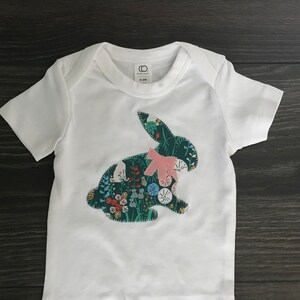 May include: White baby onesie with a green and floral bunny applique. The bunny has a pink bow.