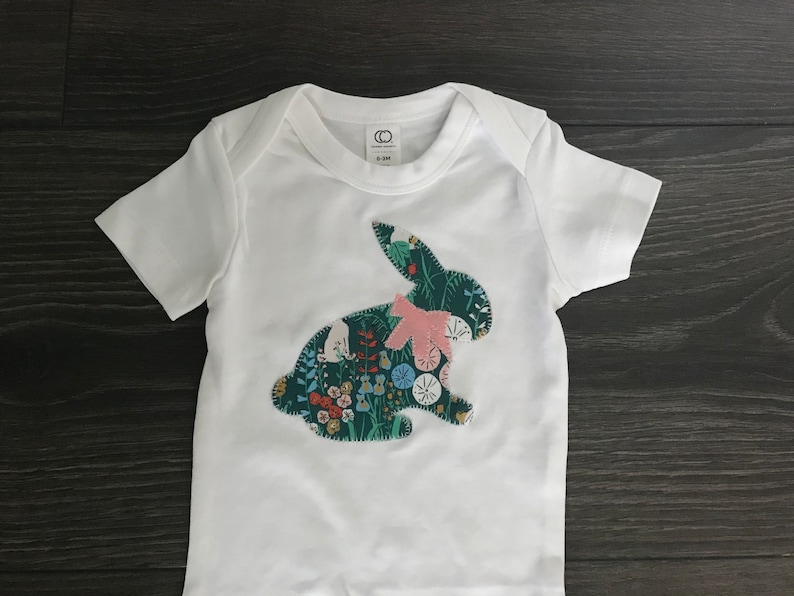 May include: White baby onesie with a green and floral bunny applique. The bunny has a pink bow.