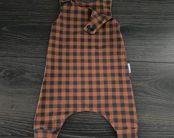 May include: A sleeveless romper featuring a brown and black buffalo check pattern. The garment has a rounded neckline, a small overlapping front flap secured with two dark buttons, and additional buttons on the shoulder straps with cuffed leg openings.