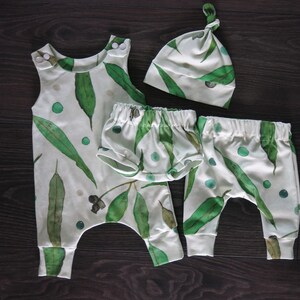May include: A baby clothing set featuring a white background with a green eucalyptus leaf pattern. The set includes a sleeveless romper, shorts, pants, and a knotted cap. The romper has snap closures at the shoulders. The shorts and pants have elastic waistbands.