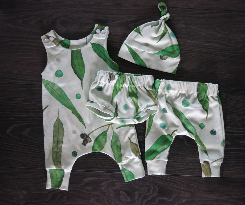 May include: A baby clothing set featuring a white background with a green eucalyptus leaf pattern. The set includes a sleeveless romper, shorts, pants, and a knotted cap. The romper has snap closures at the shoulders. The shorts and pants have elastic waistbands.