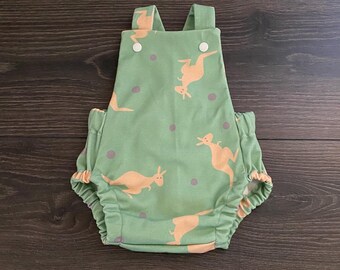 May include: A sage green romper featuring a repeating pattern of light orange kangaroos and small grey dots. The garment has two shoulder straps with white snap buttons at the front and elasticized leg openings for a gathered, comfortable fit.