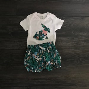 May include: A white bodysuit with a pink and green bunny applique. The bodysuit is paired with green and white floral print bloomers.