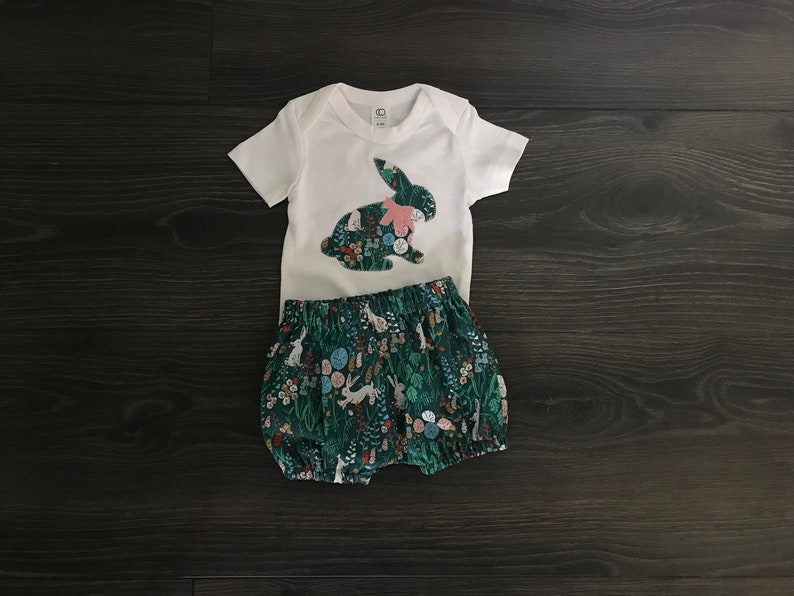 May include: A white bodysuit with a pink and green bunny applique. The bodysuit is paired with green and white floral print bloomers.