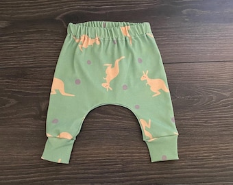 May include: Light green pants featuring a repeating pattern of light orange kangaroos and small grey dots. The pants have an elasticized waistband and fitted cuffs at the ankles, designed with a drop-crotch silhouette for a relaxed fit.