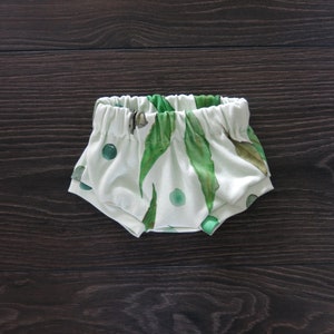 May include: White baby bloomers with an elastic waistband, featuring a watercolor print of green leaves and circular accents. The fabric is soft and the design is gender-neutral, suitable for infants.