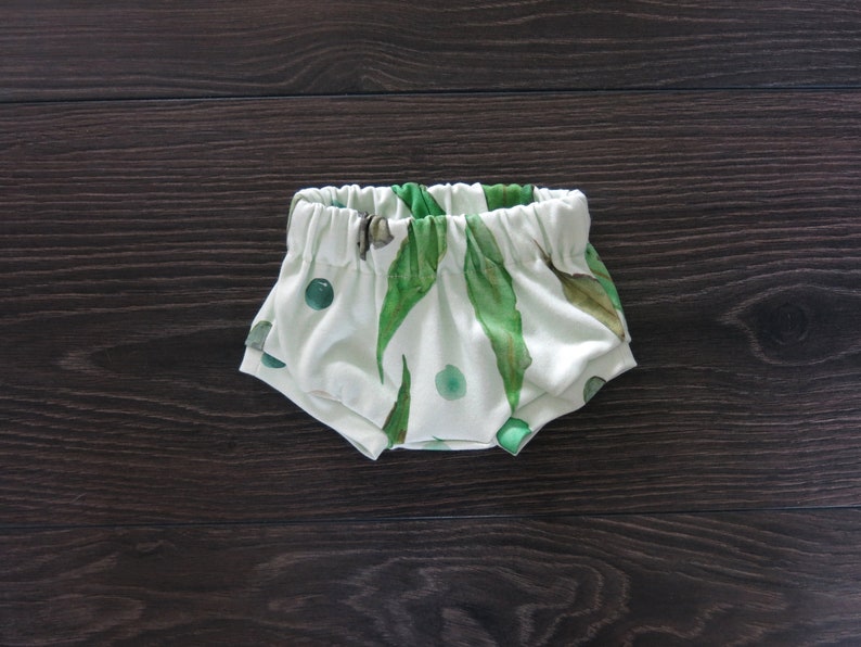 May include: White baby bloomers with an elastic waistband, featuring a watercolor print of green leaves and circular accents. The fabric is soft and the design is gender-neutral, suitable for infants.