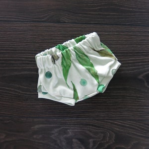 May include: White baby shorts with an elastic waistband, featuring a green leaf and dot pattern. The shorts are made of soft fabric and are designed for comfort. The overall theme is baby clothing.