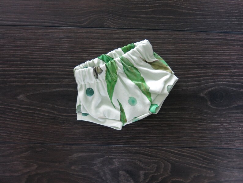 May include: White baby shorts with an elastic waistband, featuring a green leaf and dot pattern. The shorts are made of soft fabric and are designed for comfort. The overall theme is baby clothing.