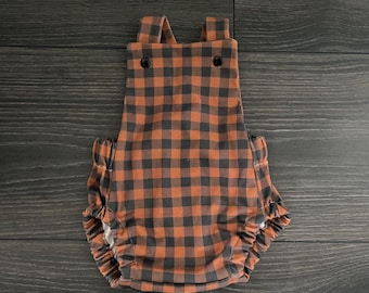May include: A sleeveless romper featuring a brown and black buffalo check pattern. The garment has two shoulder straps with dark buttons at the front chest and elasticized leg openings with gathered fabric details along the sides.