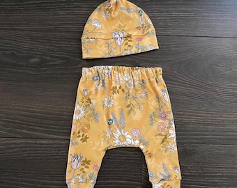 May include: A mustard yellow baby outfit consisting of a knotted beanie hat and matching pants. Both pieces feature a repeating floral print with white, pink, blue, and green flowers and foliage. The pants have an elastic waistband and cuffs.
