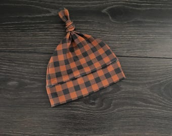 May include: A baby hat featuring a burnt orange and charcoal gray buffalo plaid pattern. The hat has a folded brim and a knotted top detail, constructed from a soft fabric with a consistent checkered design throughout the entire garment.