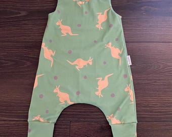 May include: A sleeveless sage green romper featuring a repeating pattern of light orange kangaroos and small purple dots. The garment has two white snap buttons on each shoulder strap and cuffed leg openings, with a small white brand tag on the side.