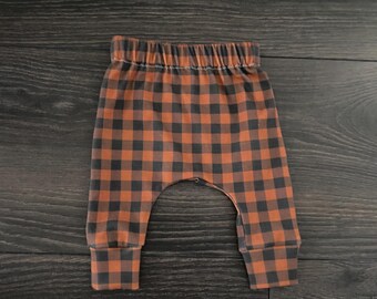May include: A pair of pants featuring a dark charcoal gray and burnt orange buffalo plaid pattern. The garment has an elasticized waistband and fitted cuffs at the ankles, designed with a drop-crotch silhouette for a relaxed fit.