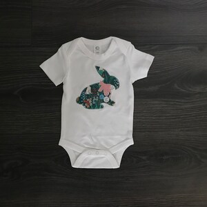 May include: White baby onesie with a green and pink floral bunny applique.