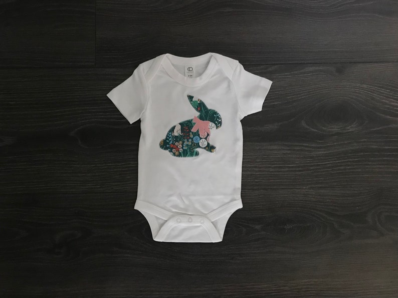 May include: White baby onesie with a green and pink floral bunny applique.