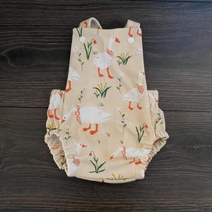 May include: A beige baby romper featuring a pattern of white geese with red feet and beaks, wearing polka dot neck scarves. Green grass and white flowers are also in the design. The romper has ruffled edges and button closures.