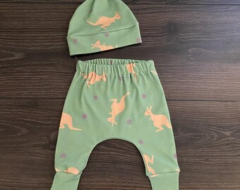 May include: A light green baby hat with a top knot and matching pants featuring a repeating pattern of light orange kangaroos and small grey dots. The pants have an elastic waistband and cuffed ankles, while the hat has a wide folded brim.