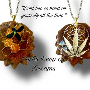 May include: Two gold necklaces with pendants made from a brown, textured material. The pendant on the left features a honeycomb design with a black and green bee. The pendant on the right features a gold leaf with a geometric design and two iridescent mushrooms. The text "Don't bee so hard on yourself all the time." is written above the necklaces. The text "Castle Keep of Dreams" is written below the necklaces.
