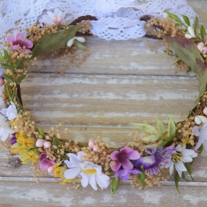 May include: A floral crown made of dried flowers and greenery. The crown is made of a variety of flowers, including white daisies, purple flowers, and pink flowers. The crown is on a wooden background.