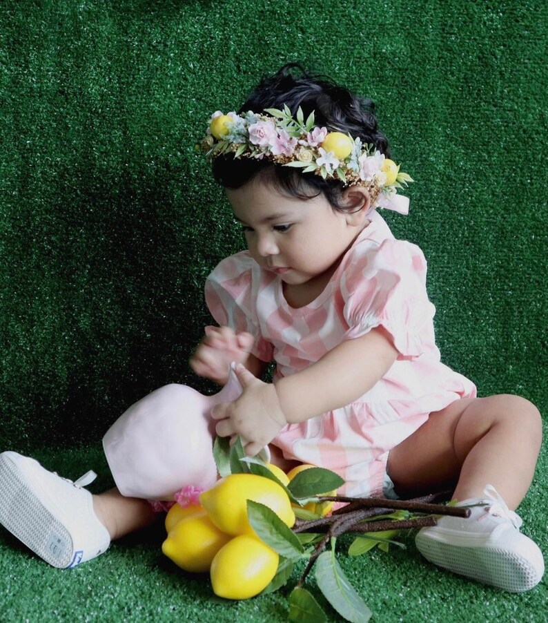 May include: A young child wearing a pink and white gingham dress and a flower crown with lemons, sitting on a green surface.