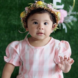 May include: A baby girl wearing a pink and white gingham dress and a flower crown with lemons. The crown has pink and yellow flowers.