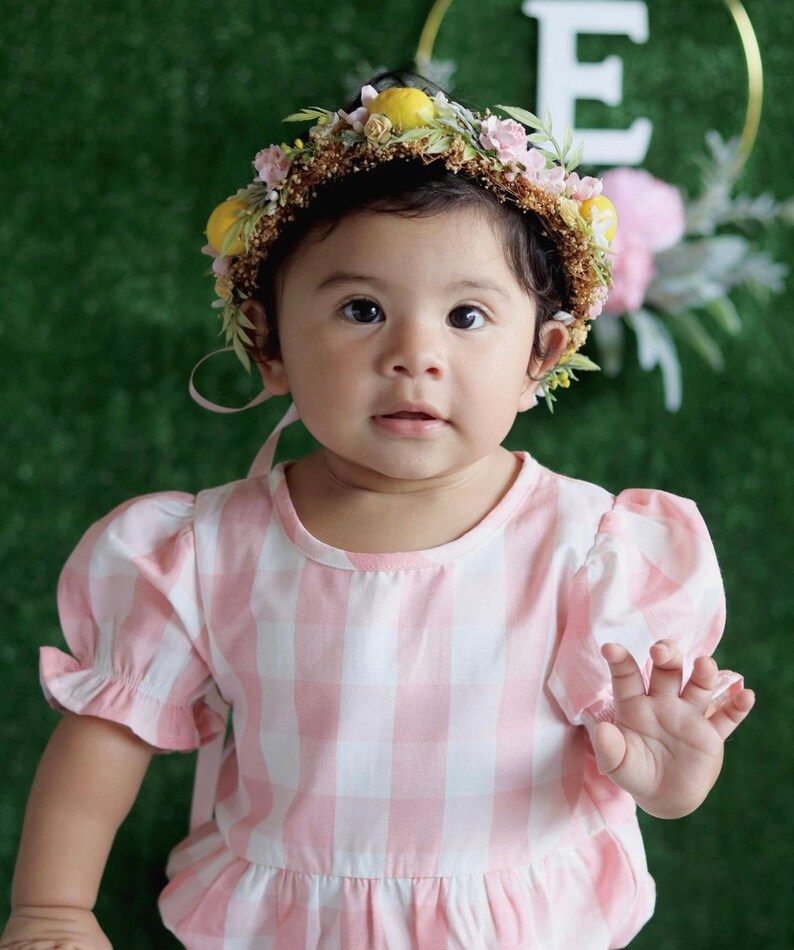 May include: A baby girl wearing a pink and white gingham dress and a flower crown with lemons. The crown has pink and yellow flowers.