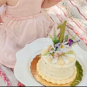 May include: A white cake with buttercream frosting decorated with flowers and a gold glitter number one candle. The cake is on a white plate with a gold rim.