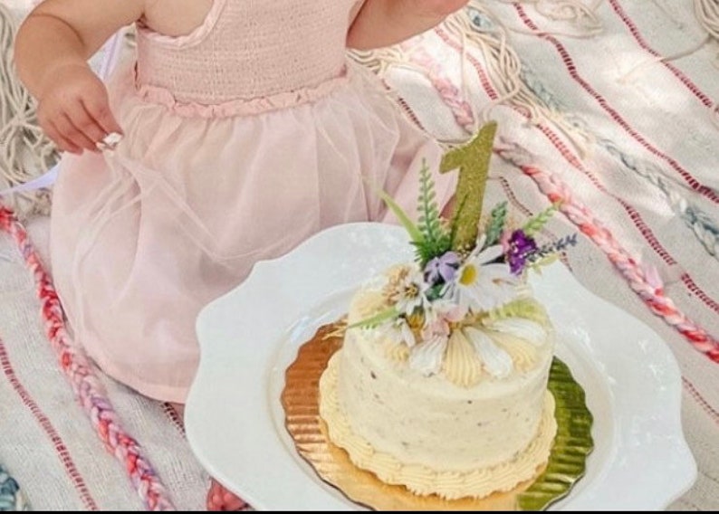 May include: A white cake with buttercream frosting decorated with flowers and a gold glitter number one candle. The cake is on a white plate with a gold rim.