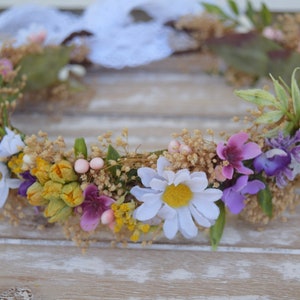May include: A floral crown with white daisies, purple and pink flowers, and yellow buds. The crown is made with dried flowers and greenery.