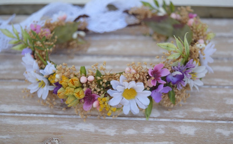 May include: A floral crown with white daisies, purple and pink flowers, and yellow buds. The crown is made with dried flowers and greenery.