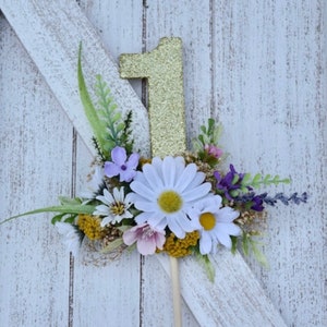 May include: Gold glitter number one cake topper with a floral arrangement of white daisies, purple flowers, and green leaves.