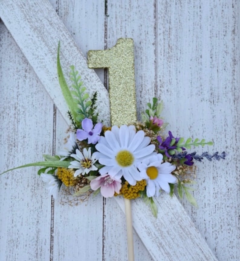 May include: Gold glitter number one cake topper with a floral arrangement of white daisies, purple flowers, and green leaves.