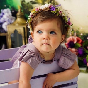 May include: A baby girl wearing a purple dress and a flower crown made of white, purple, and green flowers. She is sitting in a light purple wooden crate.
