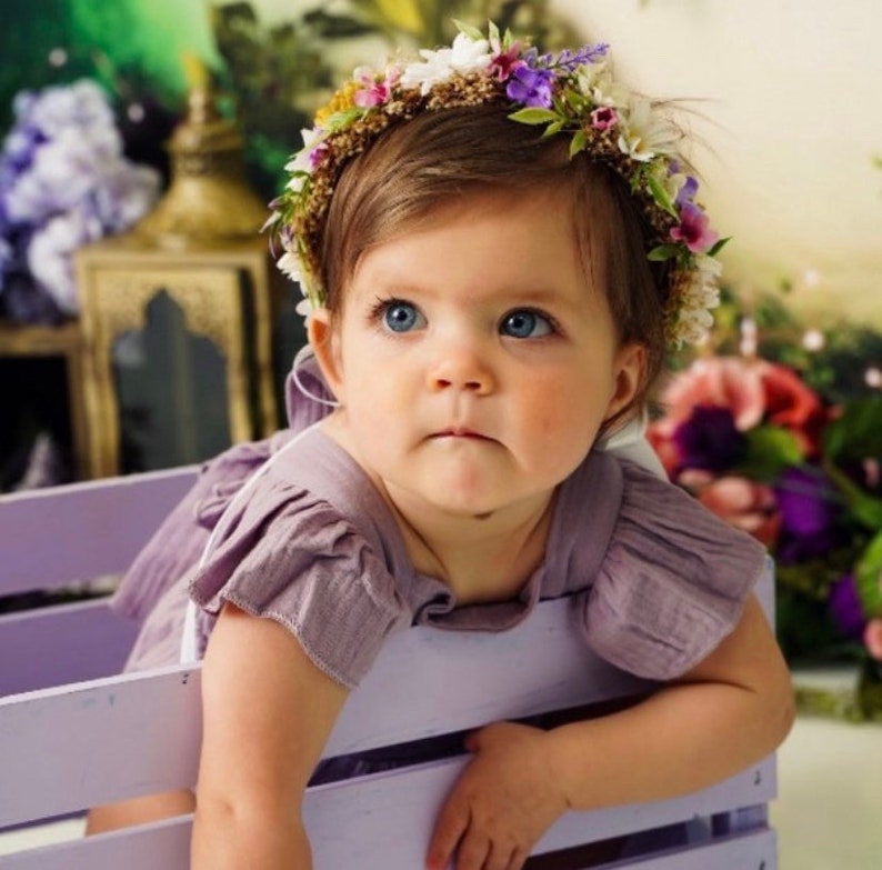 May include: A baby girl wearing a purple dress and a flower crown made of white, purple, and green flowers. She is sitting in a light purple wooden crate.
