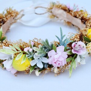 May include: A floral crown with pink, white, and yellow flowers, green leaves, and small yellow lemons. The crown is made with a brown wire base and has a white ribbon.