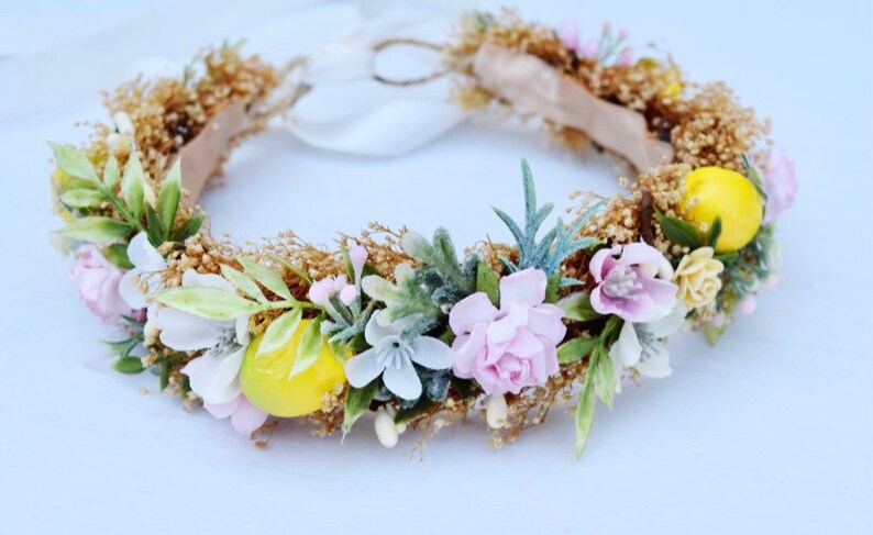 May include: A floral crown with pink, white, and yellow flowers, green leaves, and small yellow lemons. The crown is made with a brown wire base and has a white ribbon.