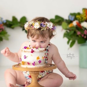 May include: A baby girl wearing a floral crown is eating a white cake decorated with colorful flowers. The cake is on a white cake stand with a wooden base.