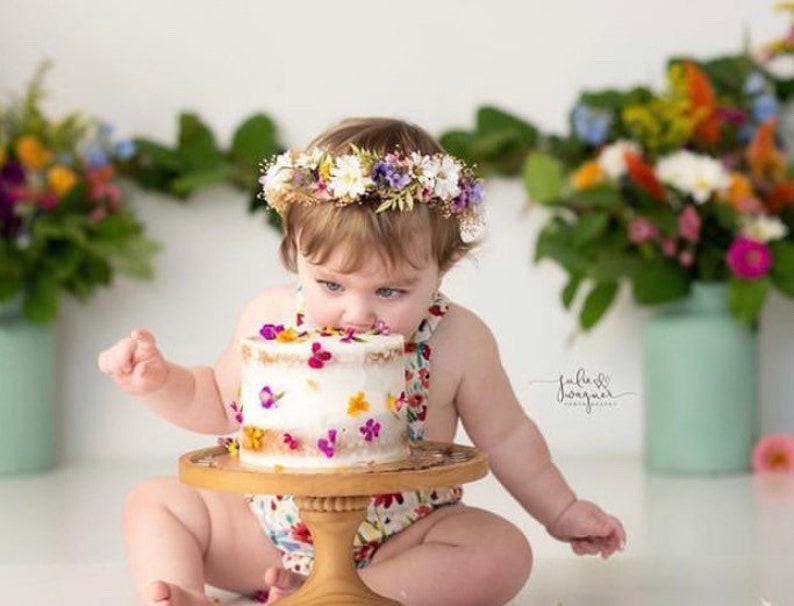 May include: A baby girl wearing a floral crown is eating a white cake decorated with colorful flowers. The cake is on a white cake stand with a wooden base.
