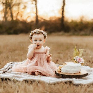 K&ouml;nnte beinhalten: Ein kleines M&auml;dchen in einem rosa Kleid und einer Blumenkrone sitzt auf einer wei&szlig;en Decke in einem Feld. Sie isst Kuchen mit ihren H&auml;nden. Ein kleiner Kuchen mit einer Kerze mit der Nummer eins steht auf einem Holzst&uuml;ck vor ihr.