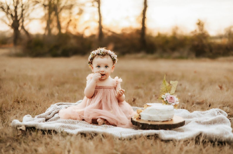 K&ouml;nnte beinhalten: Ein kleines M&auml;dchen in einem rosa Kleid und einer Blumenkrone sitzt auf einer wei&szlig;en Decke in einem Feld. Sie isst Kuchen mit ihren H&auml;nden. Ein kleiner Kuchen mit einer Kerze mit der Nummer eins steht auf einem Holzst&uuml;ck vor ihr.