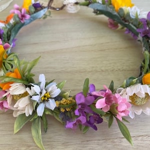 May include: A floral crown made of artificial flowers in shades of yellow, orange, white, pink, and purple. The crown is made of a variety of flowers, including daisies, roses, and wildflowers.