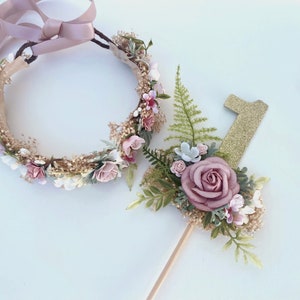 K&ouml;nnte beinhalten: Ein Blumenkranz mit einem rosa Band und einer goldenen Glitzer-Zahl eins mit floralen Akzenten. Der Kranz und der Topper sind beide mit rosa und wei&szlig;en Blumen und Gr&uuml;n verziert.
