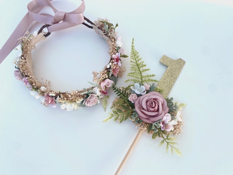 K&ouml;nnte beinhalten: Ein Blumenkranz mit einem rosa Band und einer goldenen Glitzer-Zahl eins mit floralen Akzenten. Der Kranz und der Topper sind beide mit rosa und wei&szlig;en Blumen und Gr&uuml;n verziert.